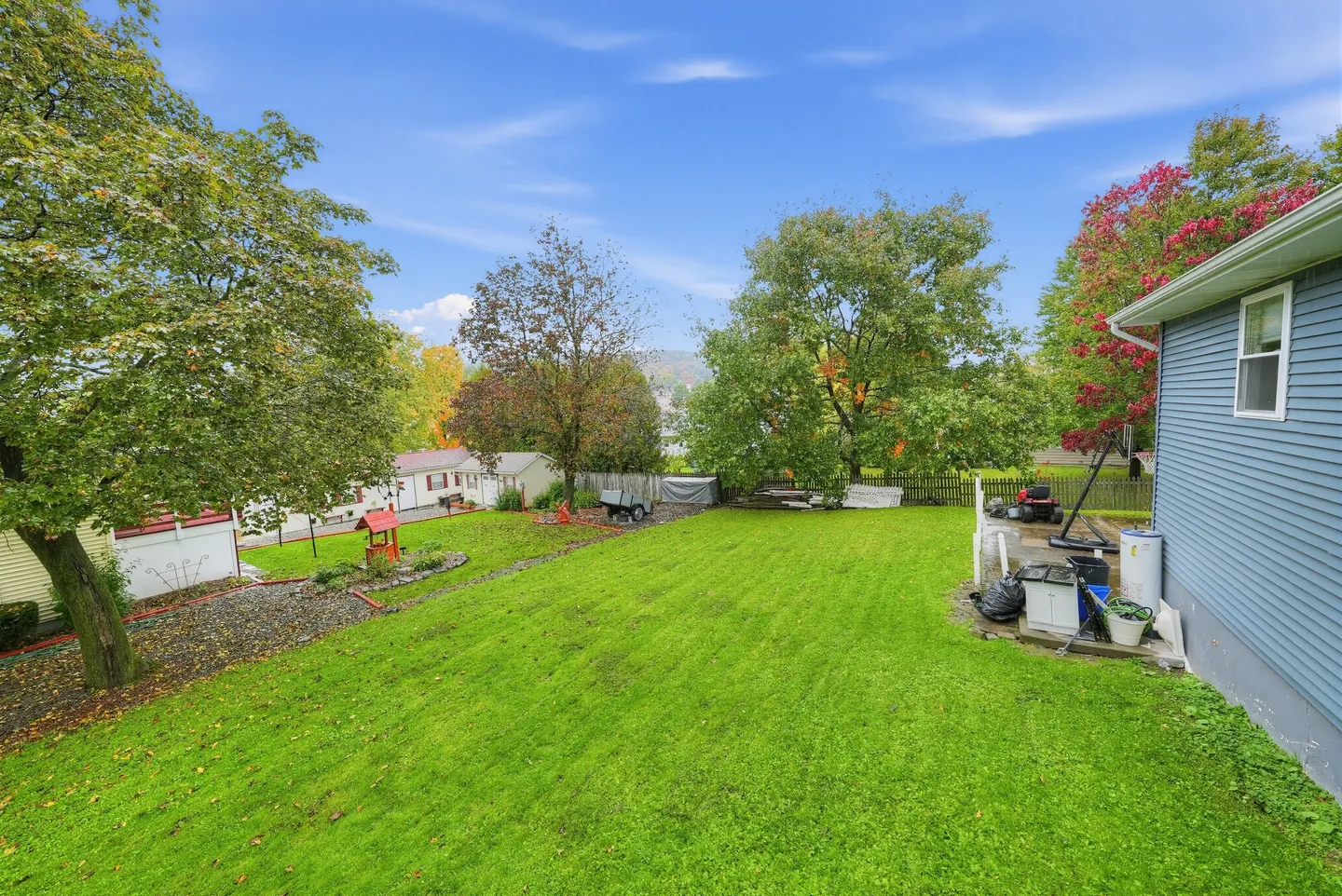 A wide, grassy backyard sloping downward with surrounding trees, a partial view of a blue-sided house on the right, and distant hills under a blue sky.