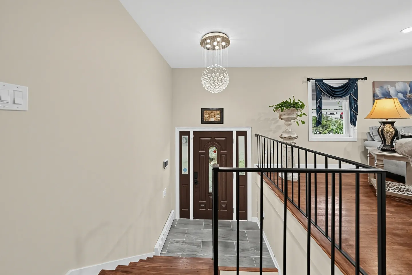 Entryway viewed from an upper landing with a dark front door, tiled landing, modern crystal light fixture, and black metal railings.