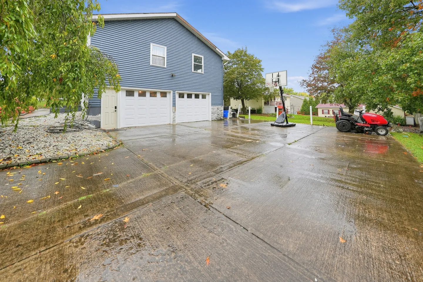Concrete driveway and two-car garage of a two-story home with blue siding, featuring a basketball hoop and riding lawnmower.