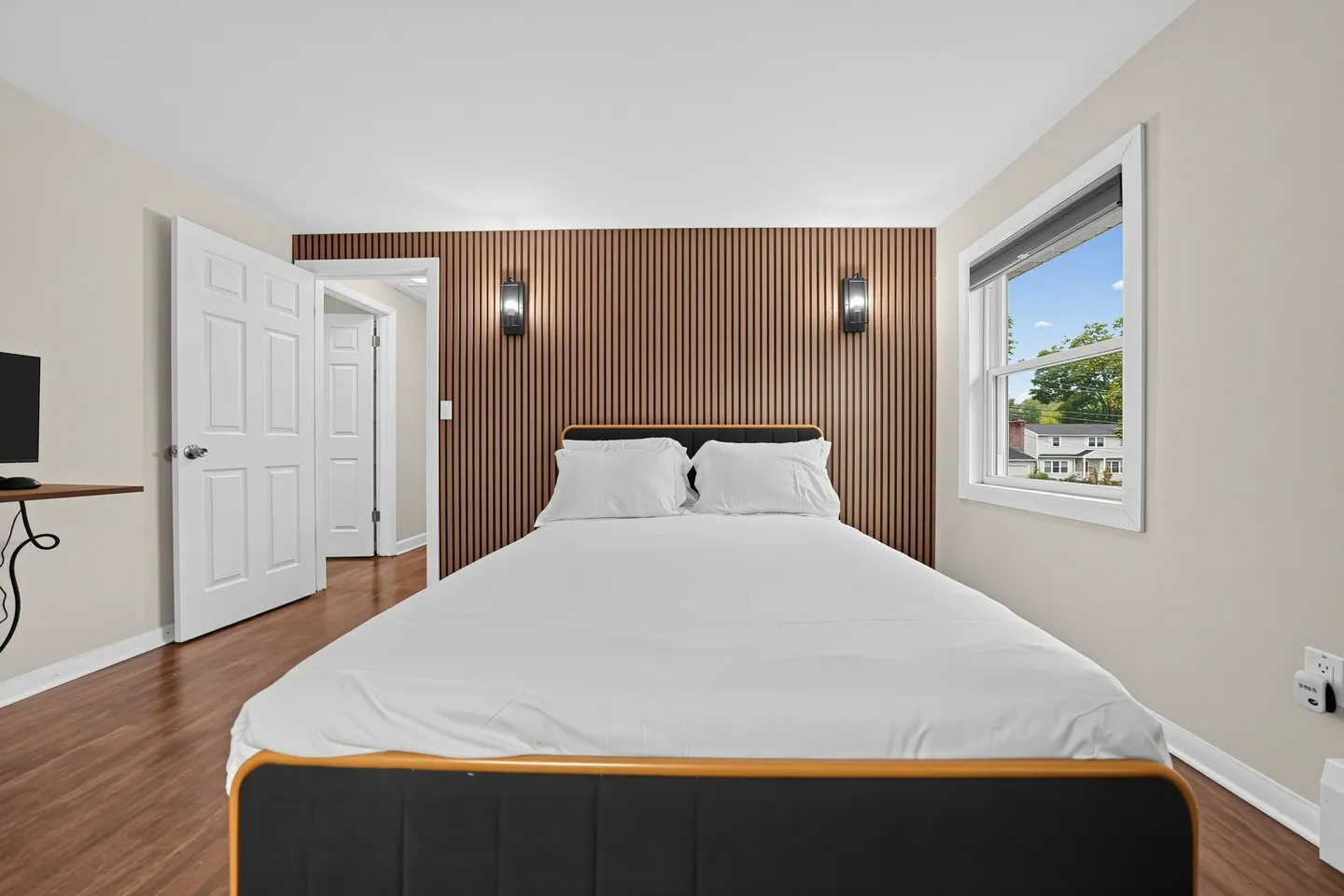 Bedroom with a bed made with white linens, a wood-slat accent wall with two mounted sconces, and a window.