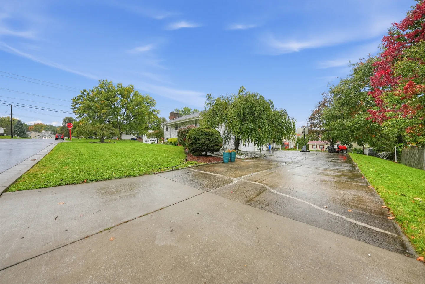 Wide concrete driveway leading past a green lawn and weeping tree toward a residential house under a blue sky.