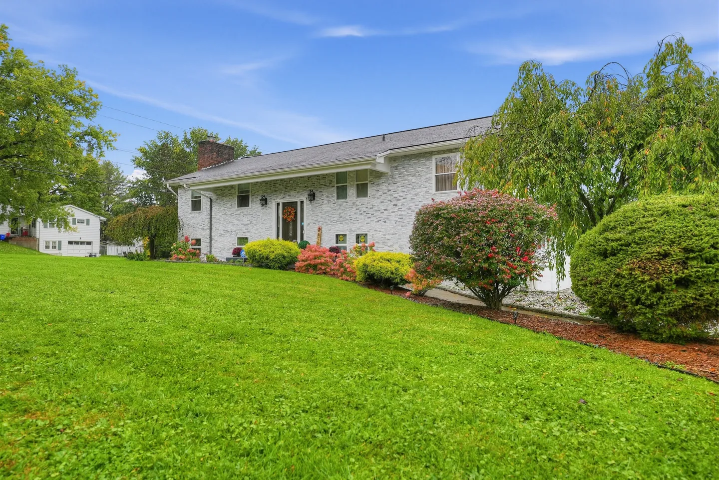 Front exterior of a white brick home on a sloping green lawn with landscaped bushes and trees.