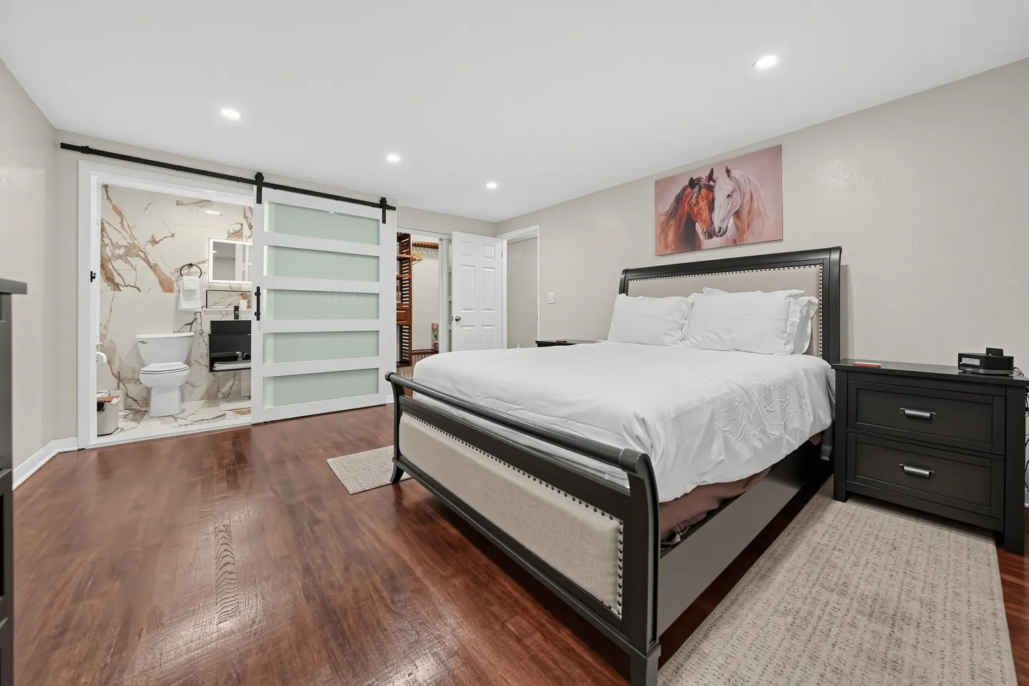 Bedroom with dark wood-style floors, a bed with an upholstered headboard, and an attached ensuite bathroom behind a sliding barn door.