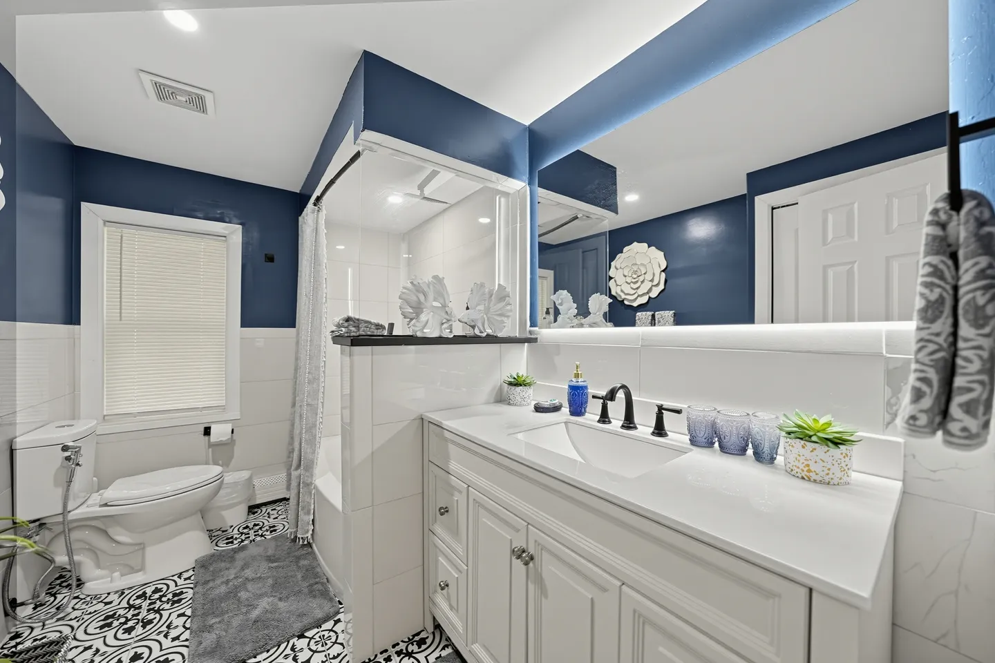 Bathroom with blue walls, patterned tile flooring, a white vanity with a large mirror, a toilet, and a tiled shower-tub combo.