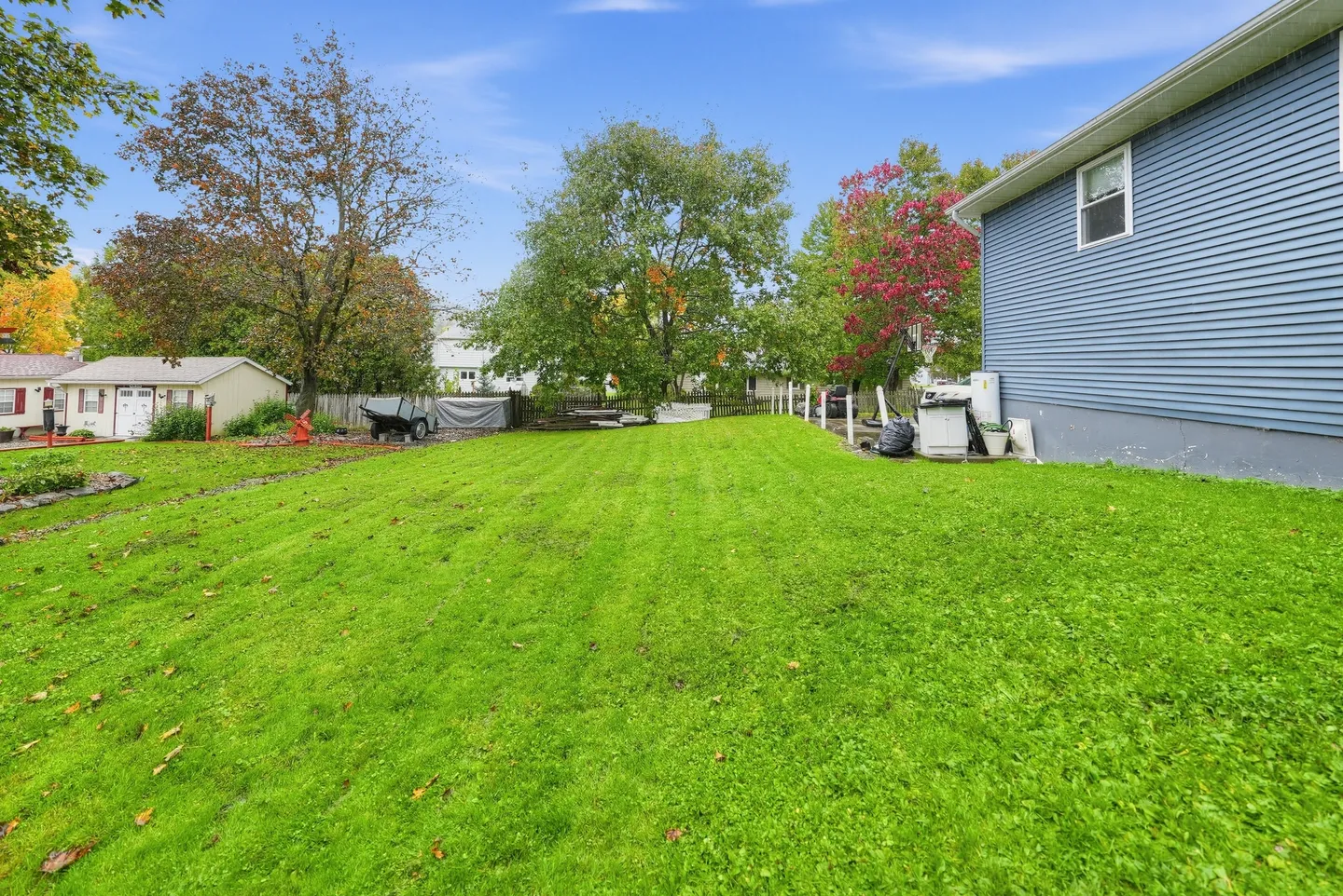 A wide, grassy backyard next to the blue horizontal siding of a house, framed by mature trees and neighboring yards under a blue sky.