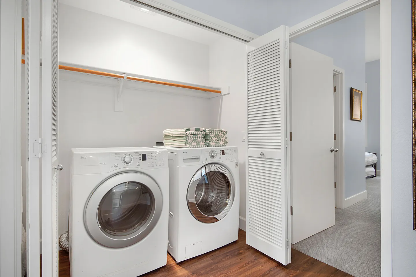 A laundry closet with white louvered bifold doors open, showing a front-loading washer and dryer with a shelf overhead.