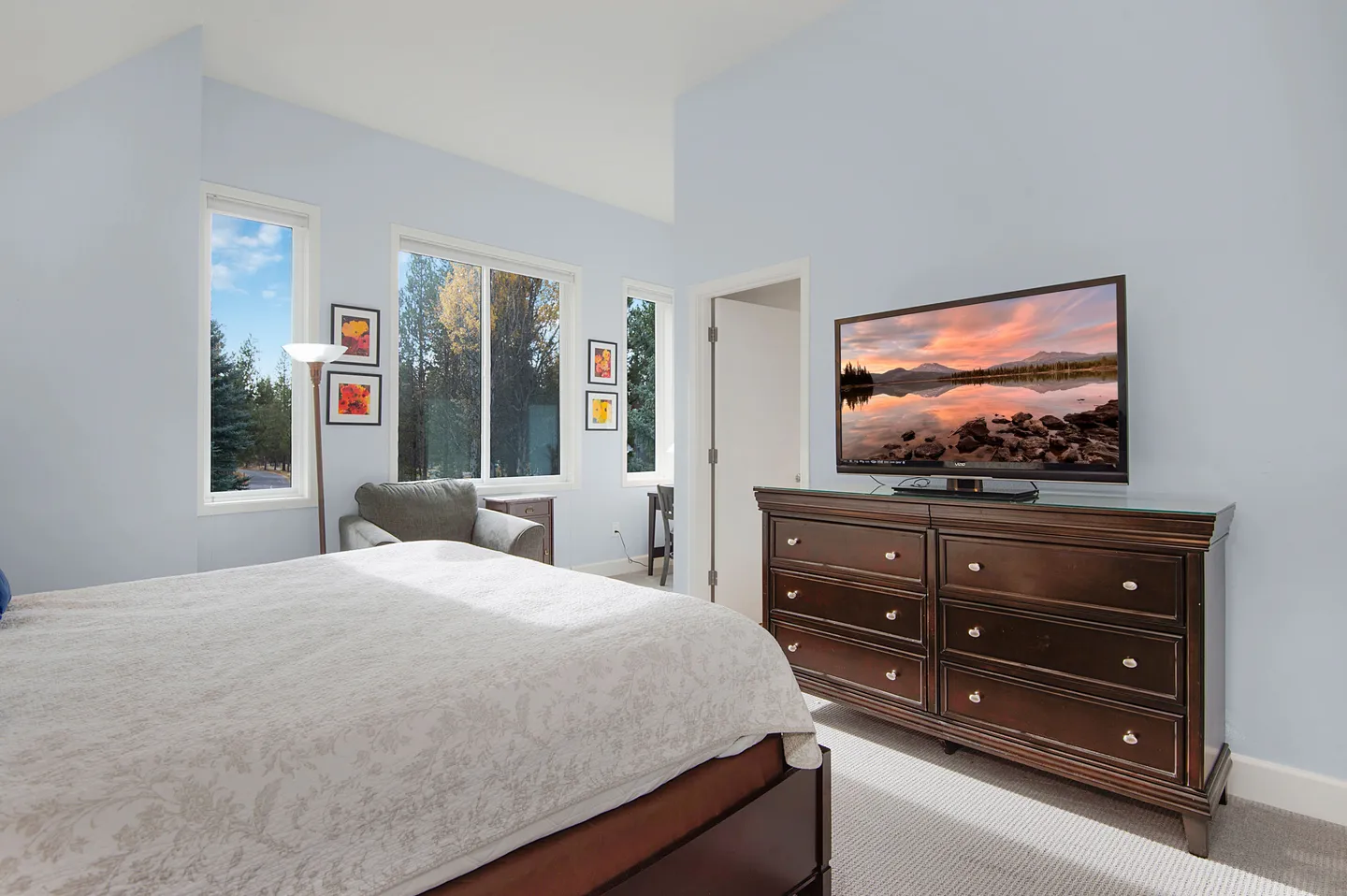 Bedroom with a bed in the foreground, a dark wood dresser with a flat-screen TV, an armchair, and windows looking out to trees.