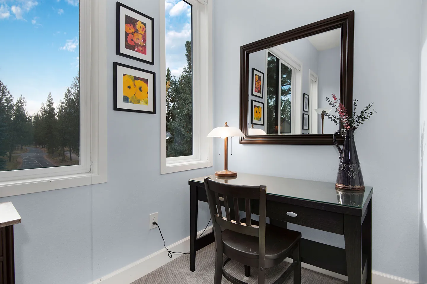 A small work nook with a dark wood desk, chair, table lamp, and large wall mirror next to tall windows.