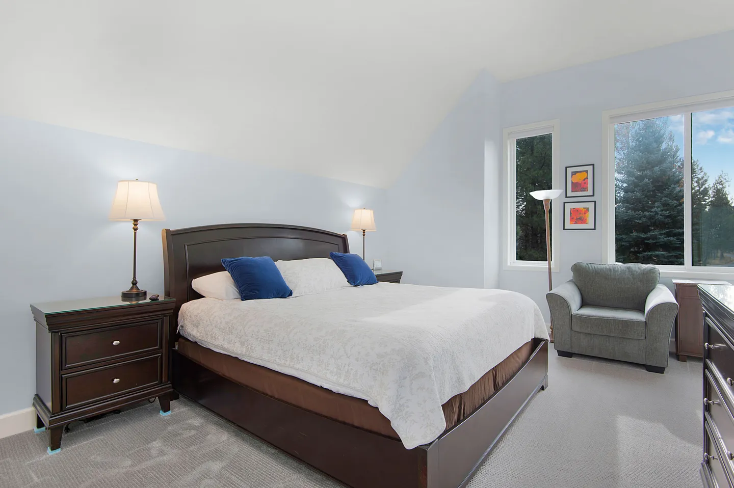 Bedroom with vaulted ceiling, large dark wood bed, matching nightstands with lamps, carpeted floor, and windows overlooking evergreen trees.