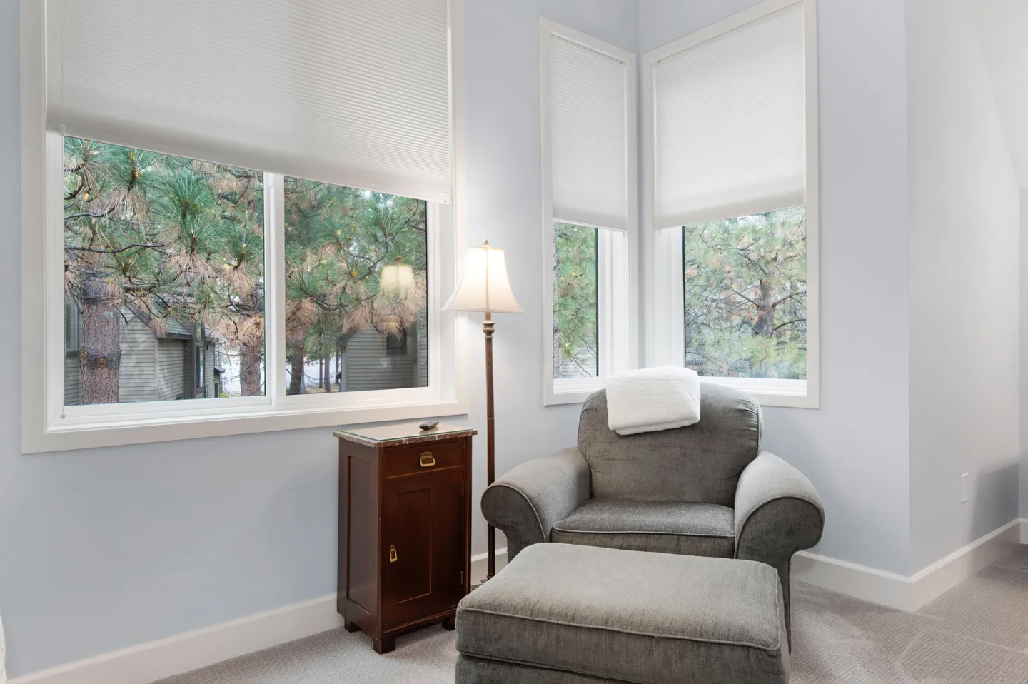 A reading nook with an upholstered grey armchair, matching ottoman, small wooden side table, floor lamp, and windows with white blinds overlooking trees.