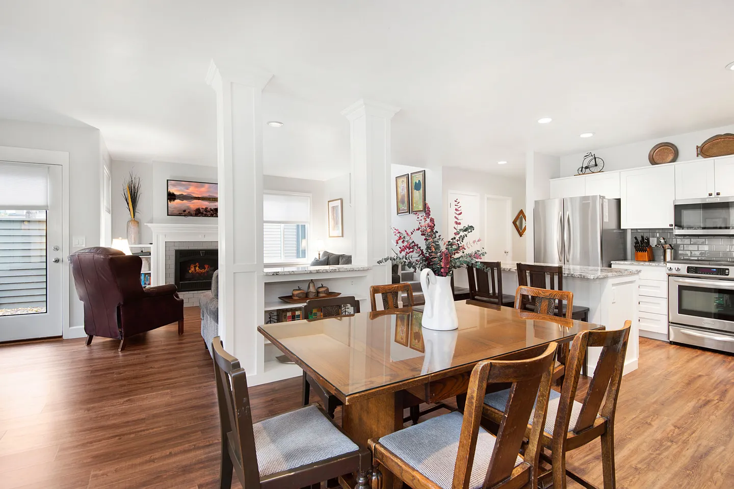 Open-concept dining area with a wooden table, leading into a living room with a fireplace and an open kitchen with white cabinets.