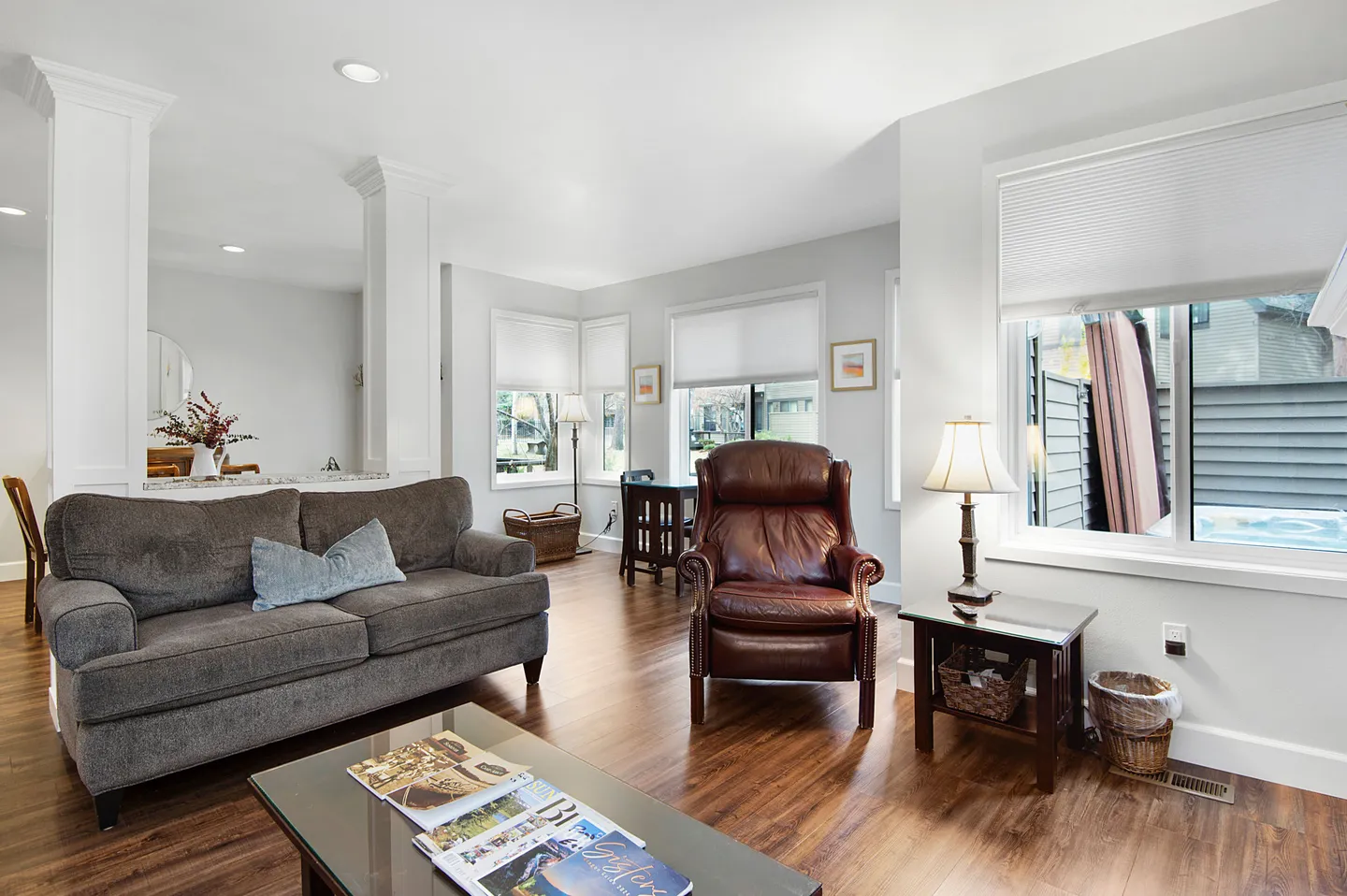 Open living area with a brown fabric sofa, a leather armchair, wood-look flooring, and windows overlooking a patio.