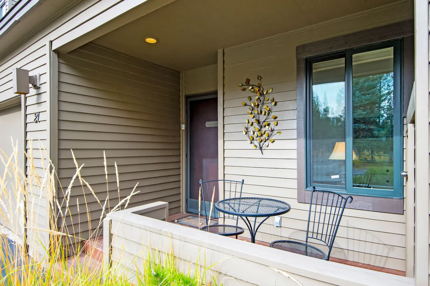 Front porch patio with siding, a small bistro table, two metal chairs, and decorative metal wall art next to a window and entrance door.