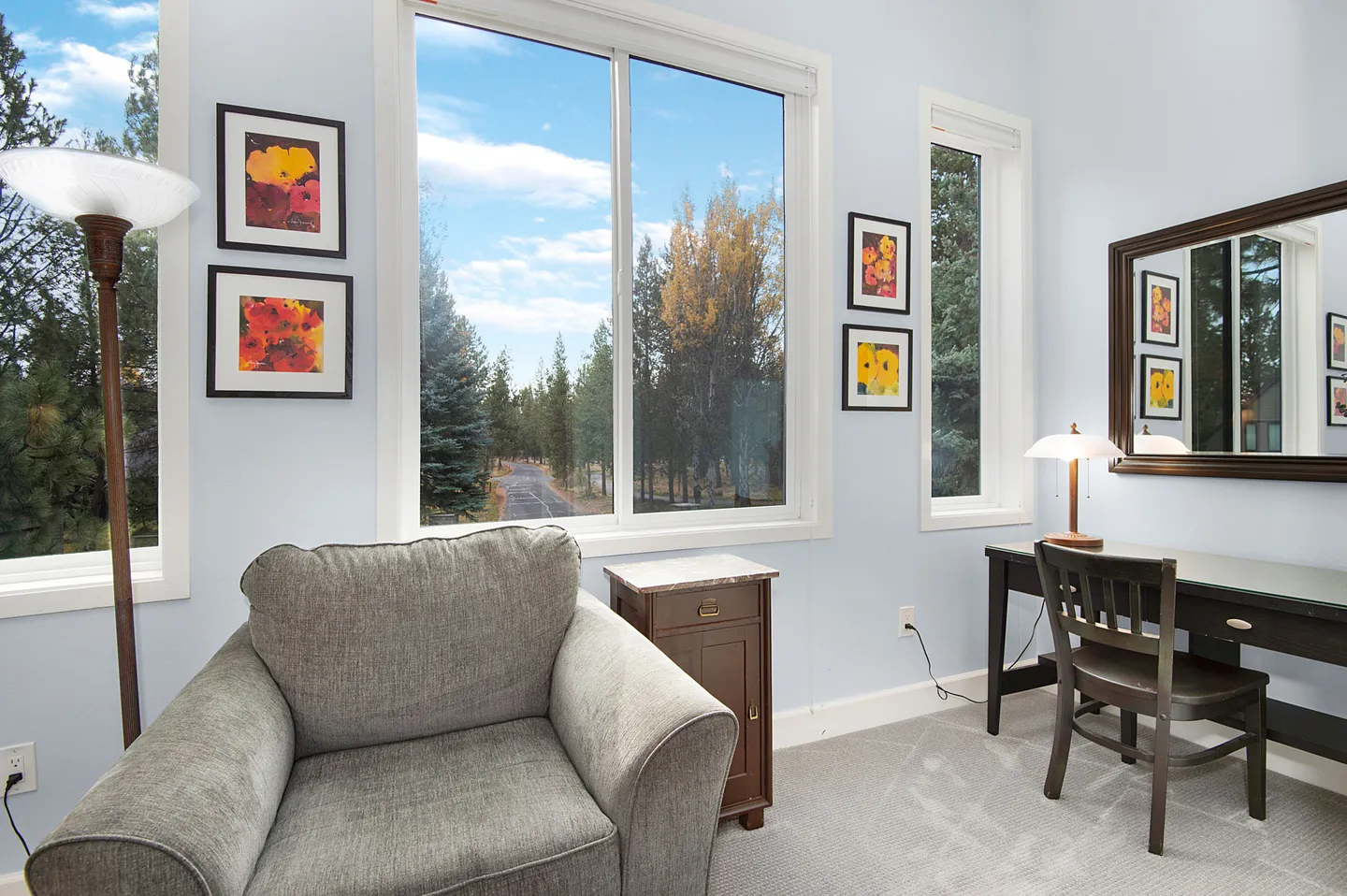 A brightly lit room featuring a gray armchair, a small work desk with a chair, and large windows looking out at pine trees.