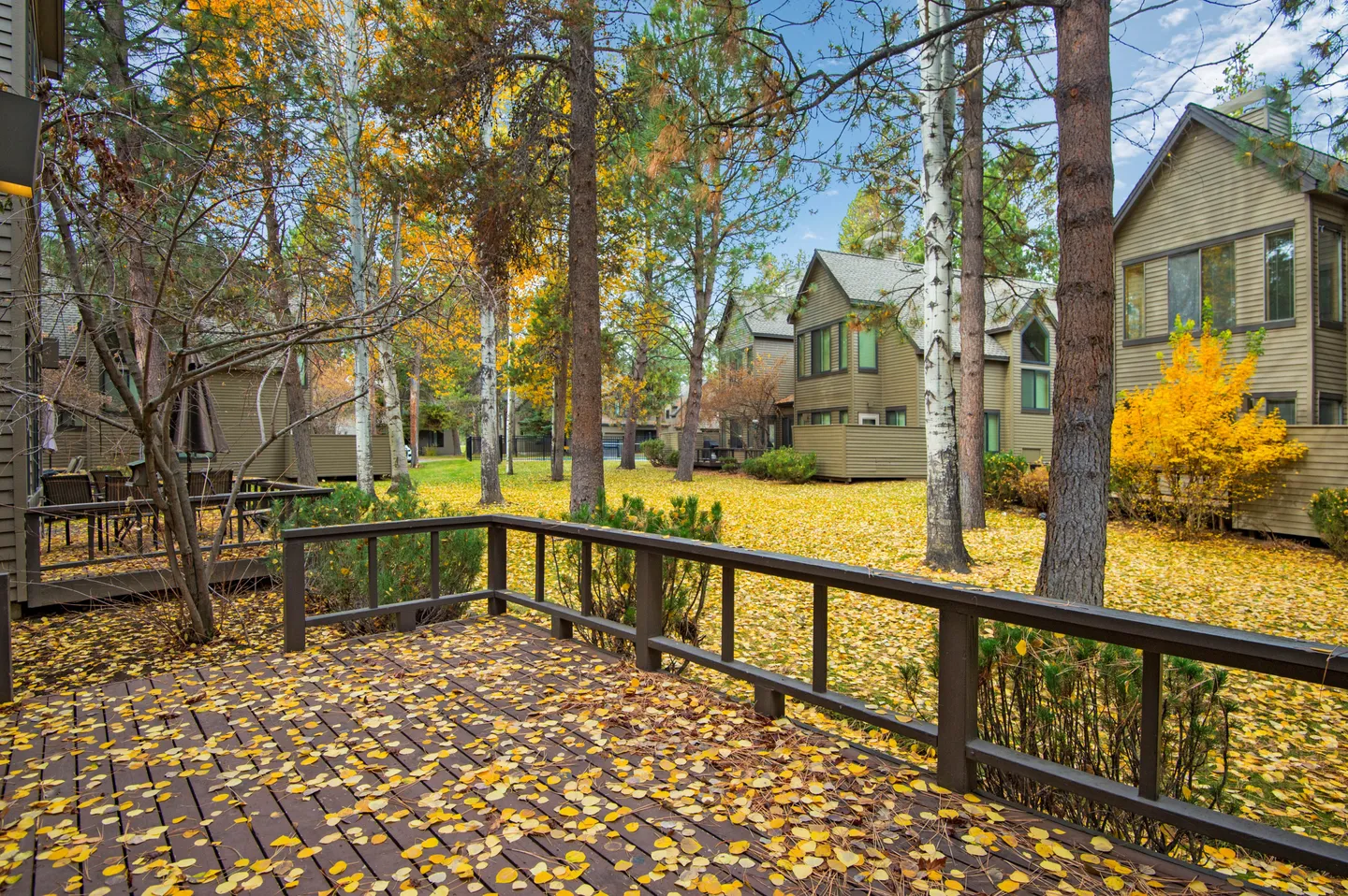 A wooden back deck scattered with yellow autumn leaves, looking out onto a wooded yard and adjacent townhomes.