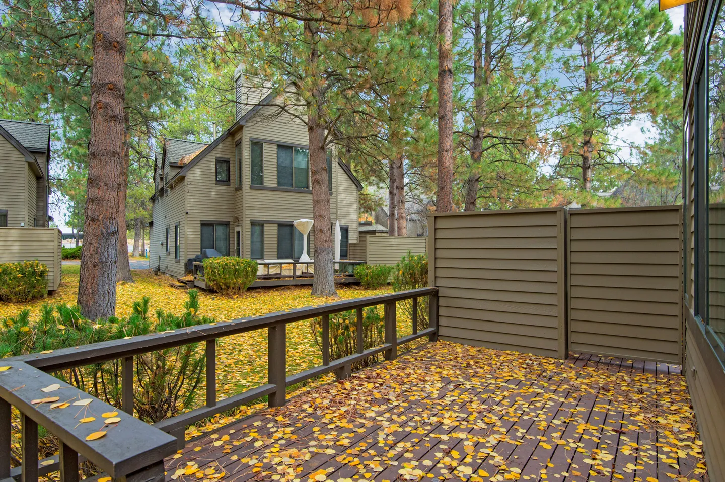 A wooden back deck scattered with yellow autumn leaves, looking out toward pine trees and neighboring townhomes.