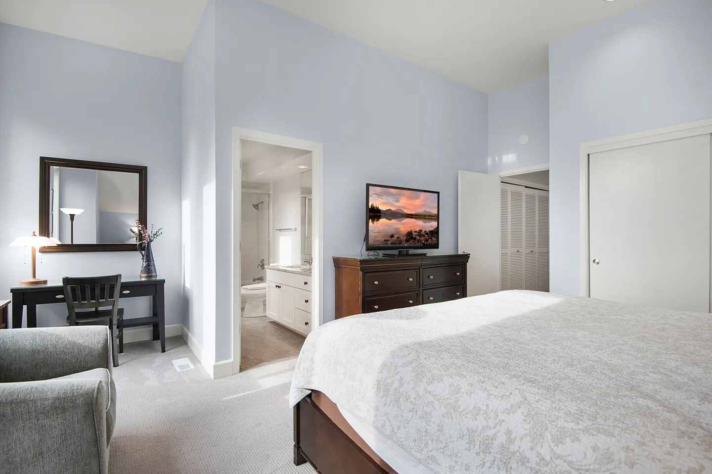 Bedroom with light blue walls, a bed in the foreground, a dark wood dresser with a TV, a desk with a mirror, and an en-suite bathroom visible through an open door.
