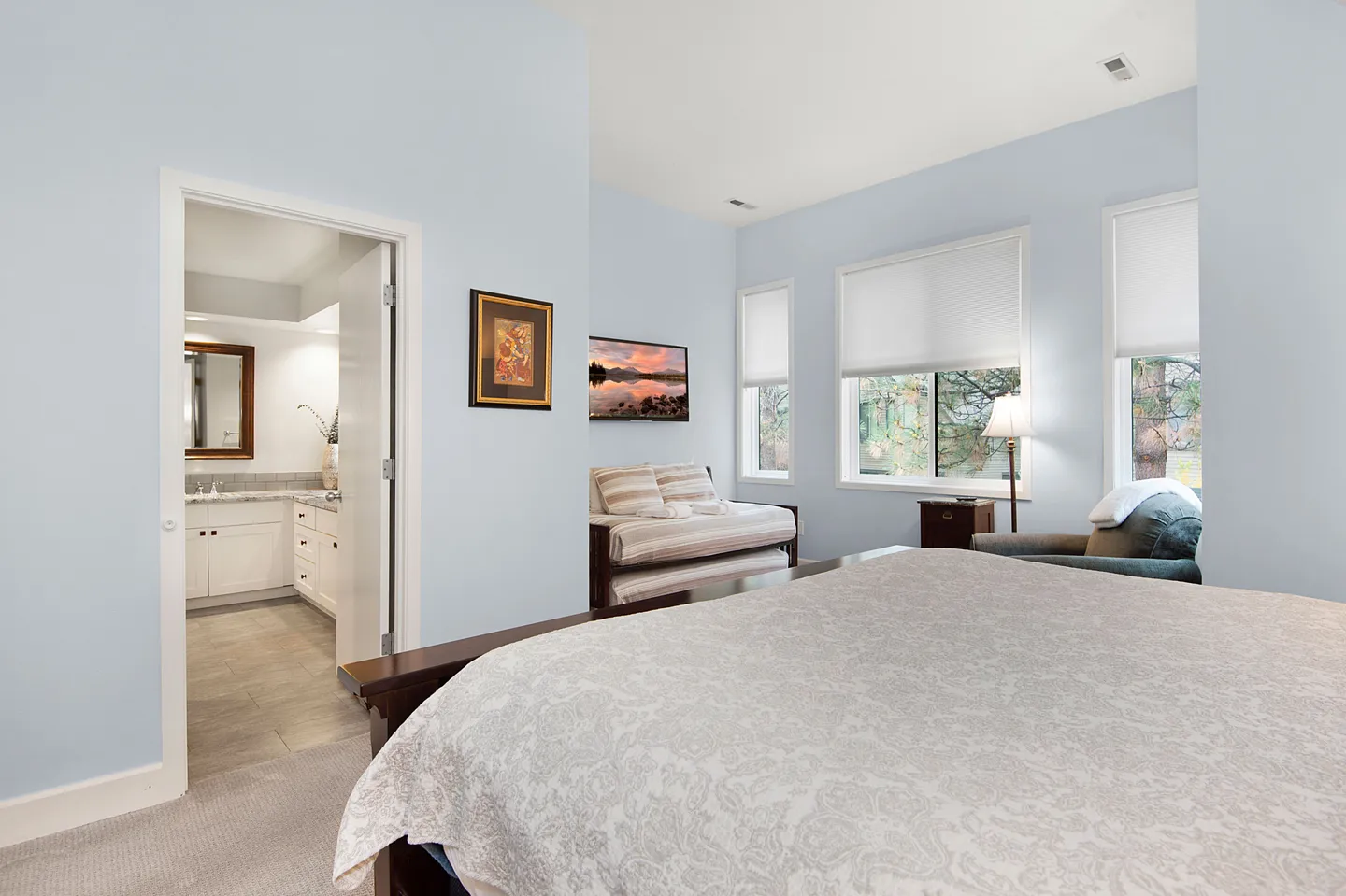 Bedroom with light blue walls, a bed in the foreground, daybed, windows, and an en-suite bathroom doorway.