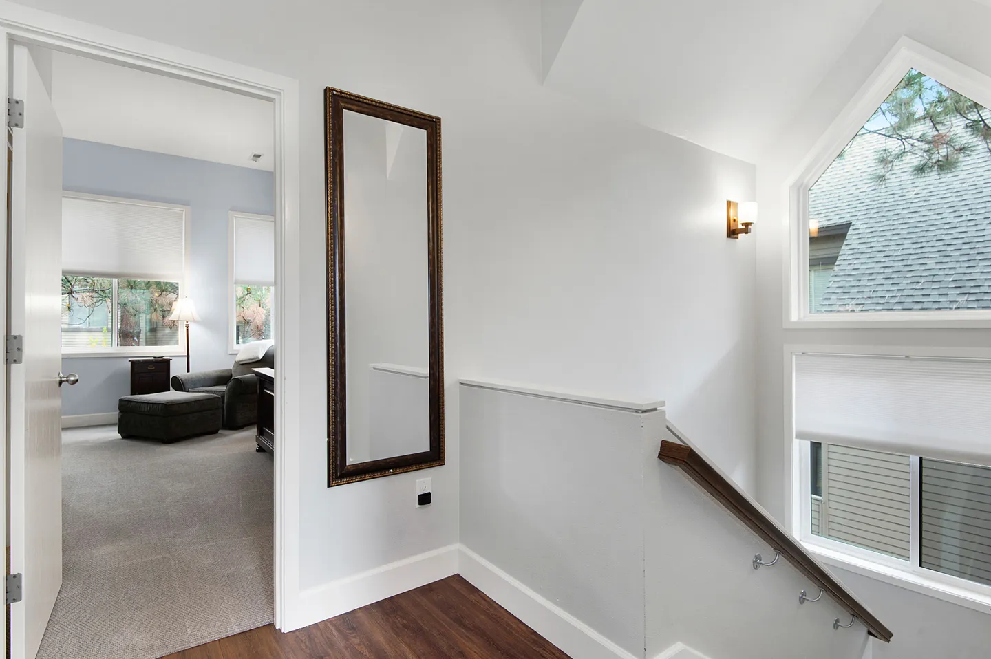 Upper-floor hallway landing next to a staircase railing with a full-length wall mirror and an open doorway leading to a carpeted room.