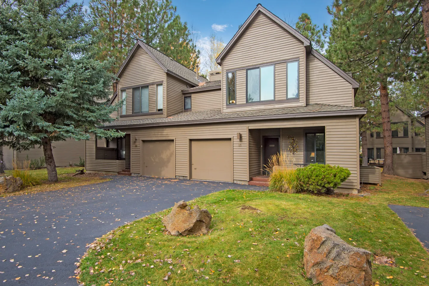 Exterior of a two-story multi-unit building with brown horizontal siding, two garage doors, and a paved driveway surrounded by pine trees and a grassy lawn with rocks.