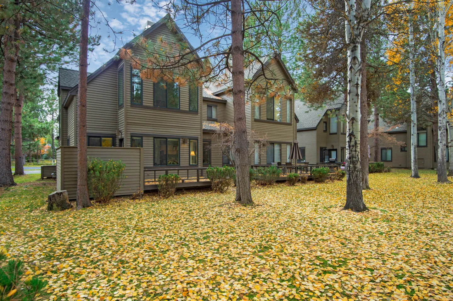 Exterior view of a multi-story home with brown siding and a back deck surrounded by trees and a lawn blanketed in yellow autumn leaves.
