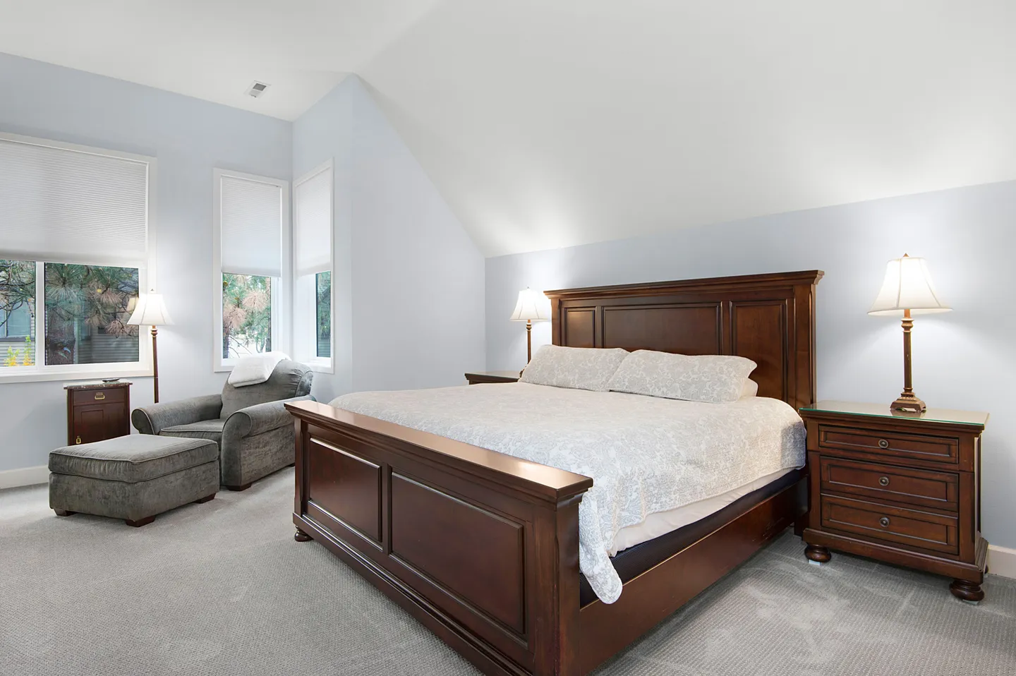 Bedroom with carpeted floor, sloped ceiling, wooden bed frame, nightstands with lamps, and an armchair with ottoman by windows.