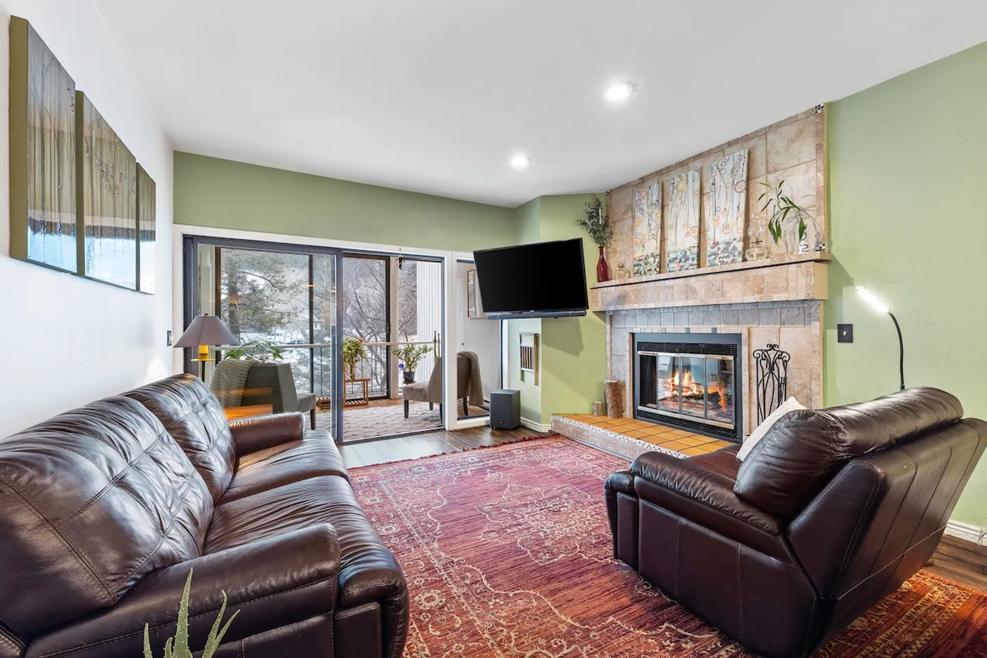 Living room with brown leather seating, a corner fireplace, mounted TV, patterned area rug, and sliding glass doors leading out to a balcony.