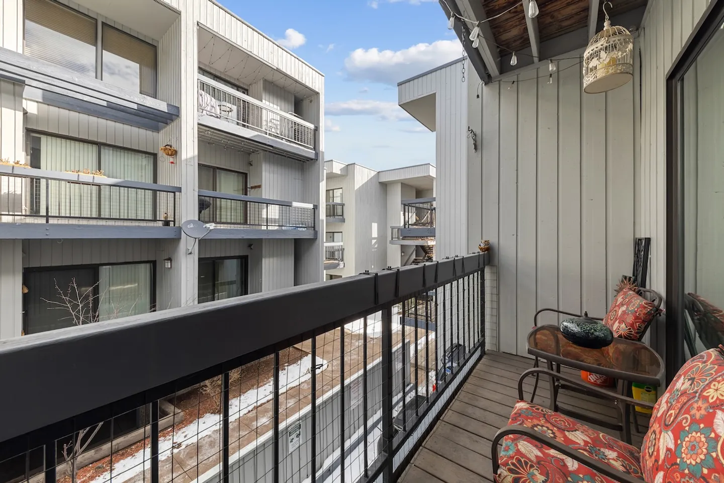 Outdoor private balcony with metal railing, two cushioned chairs, and a small table overlooking adjacent apartment buildings.