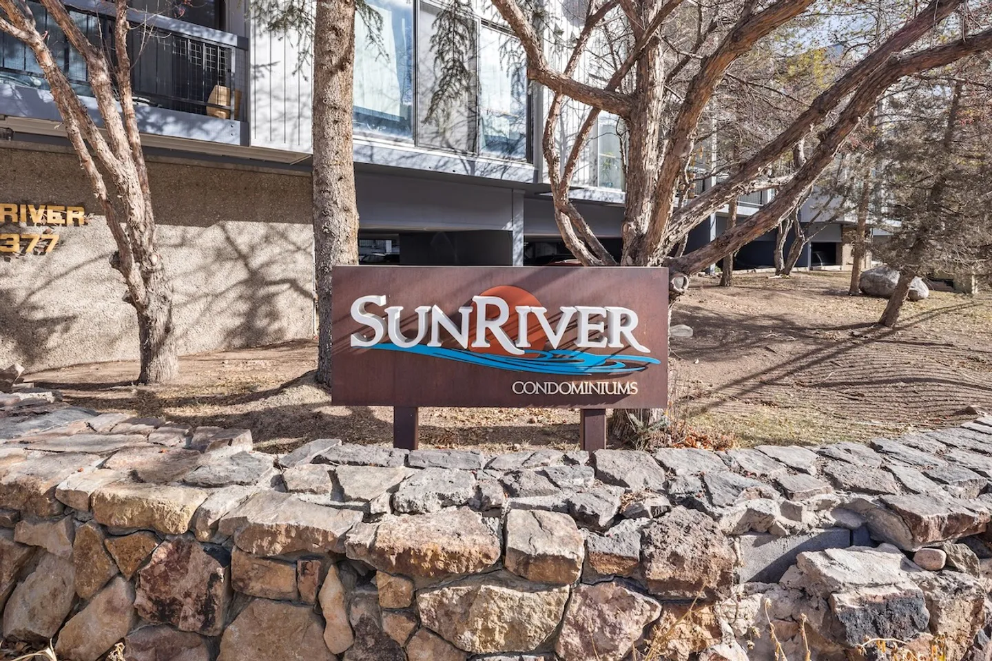 Sun River Condominiums sign set behind a low stone wall in front of a multi-story building and trees.