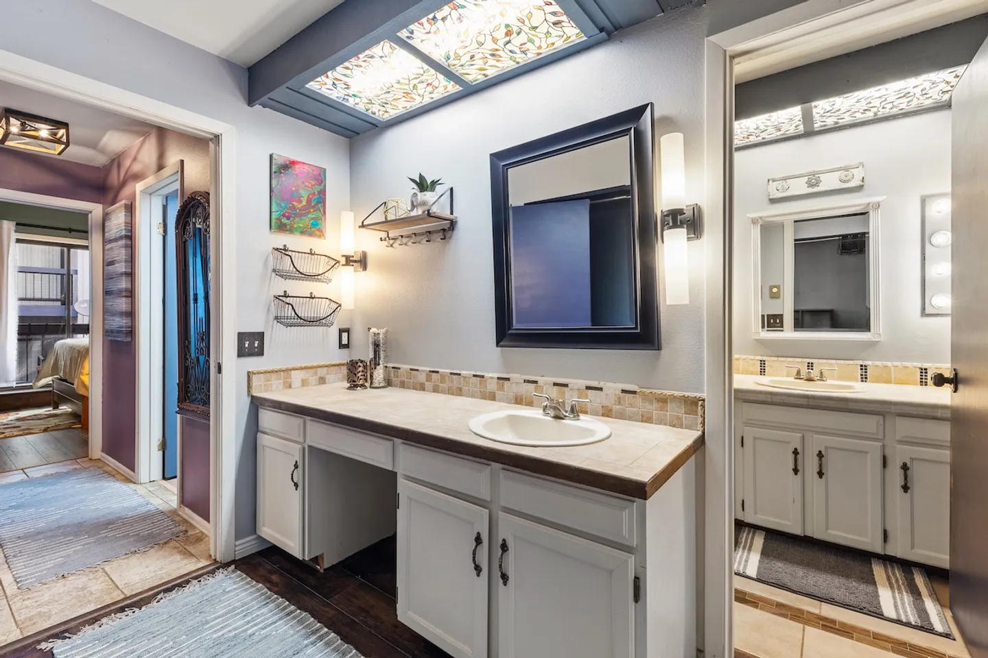 Bathroom vanity area with a tiled countertop, drop-in sink, dark framed mirror, wall sconces, and an overhead patterned light box, connecting to an adjoining bathroom.