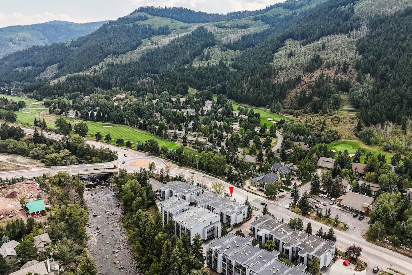 Aerial view of a residential complex with flat roofs next to a river, road, and forested mountains.