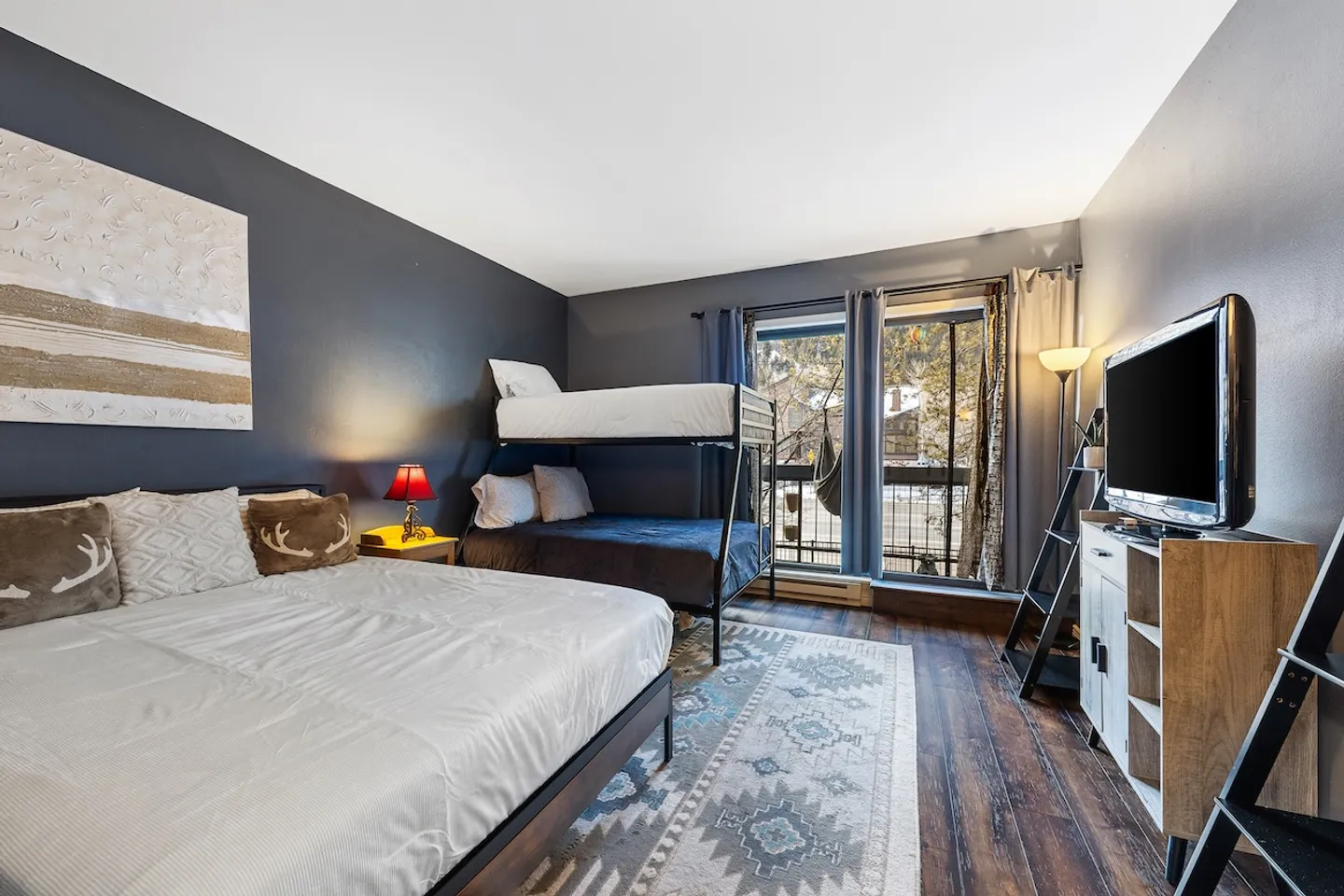 Bedroom featuring a large bed, metal bunk bed, TV console, patterned area rug, and sliding glass doors to a balcony.