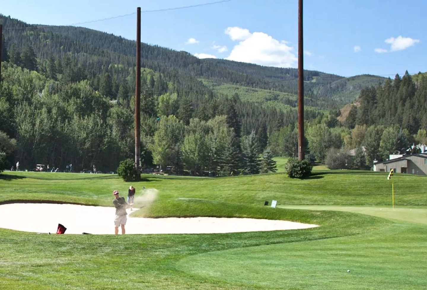 A golf course green with a player hitting a ball out of a sand bunker against a backdrop of forested mountains under a clear sky.