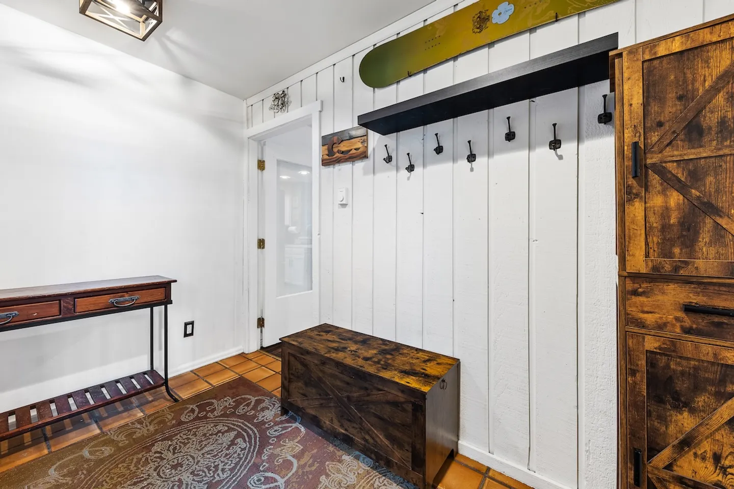 Entryway with vertical white paneled wall, storage bench, coat hooks, a narrow console table, and tile flooring with an area rug.
