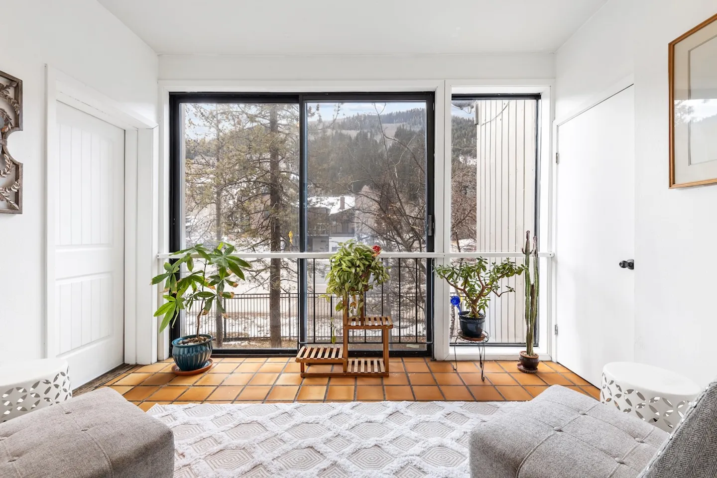 Sunlit alcove with terracotta floor tiles, potted houseplants, and sliding glass doors revealing winter mountain scenery.