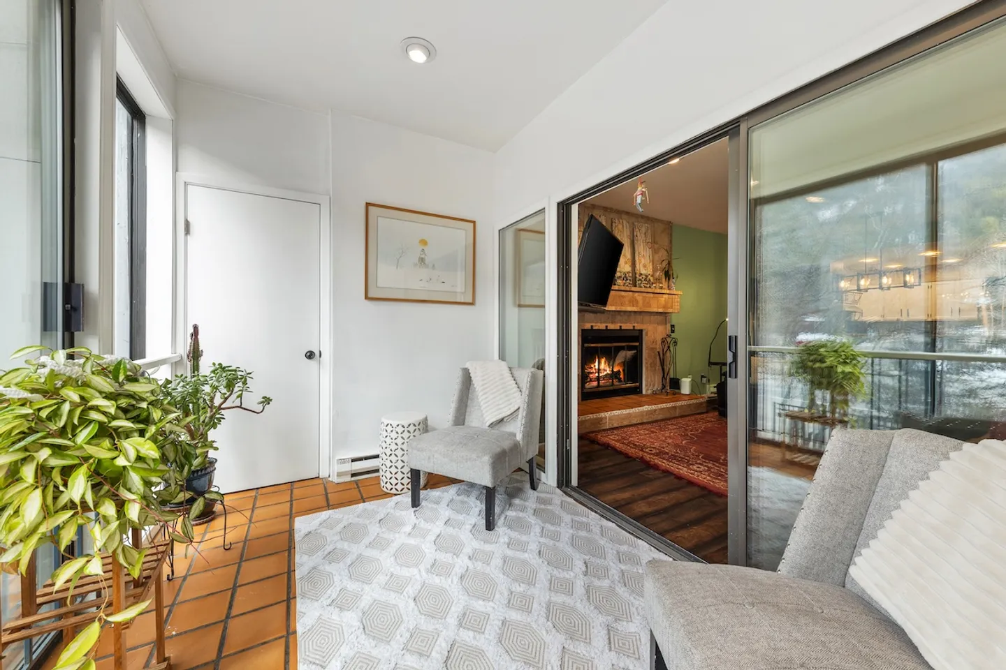 Sunroom with tiled floor, patterned area rug, armchairs, and sliding glass doors looking into a living room with a lit fireplace.