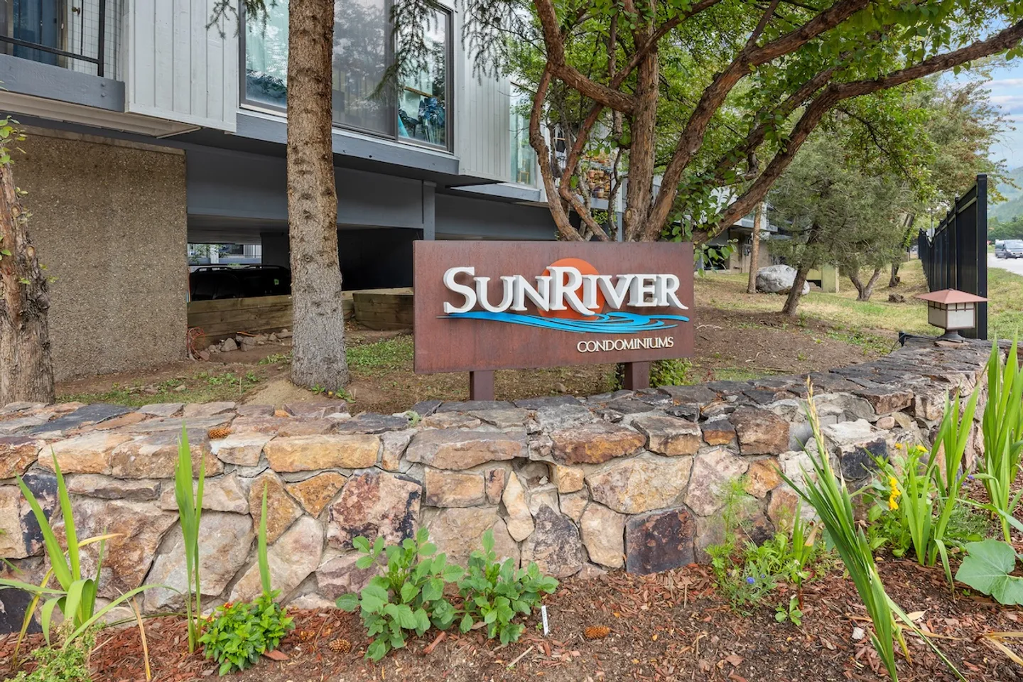 Sunriver Condominiums entrance sign set behind a curved stone retaining wall with surrounding landscaping and building exterior.