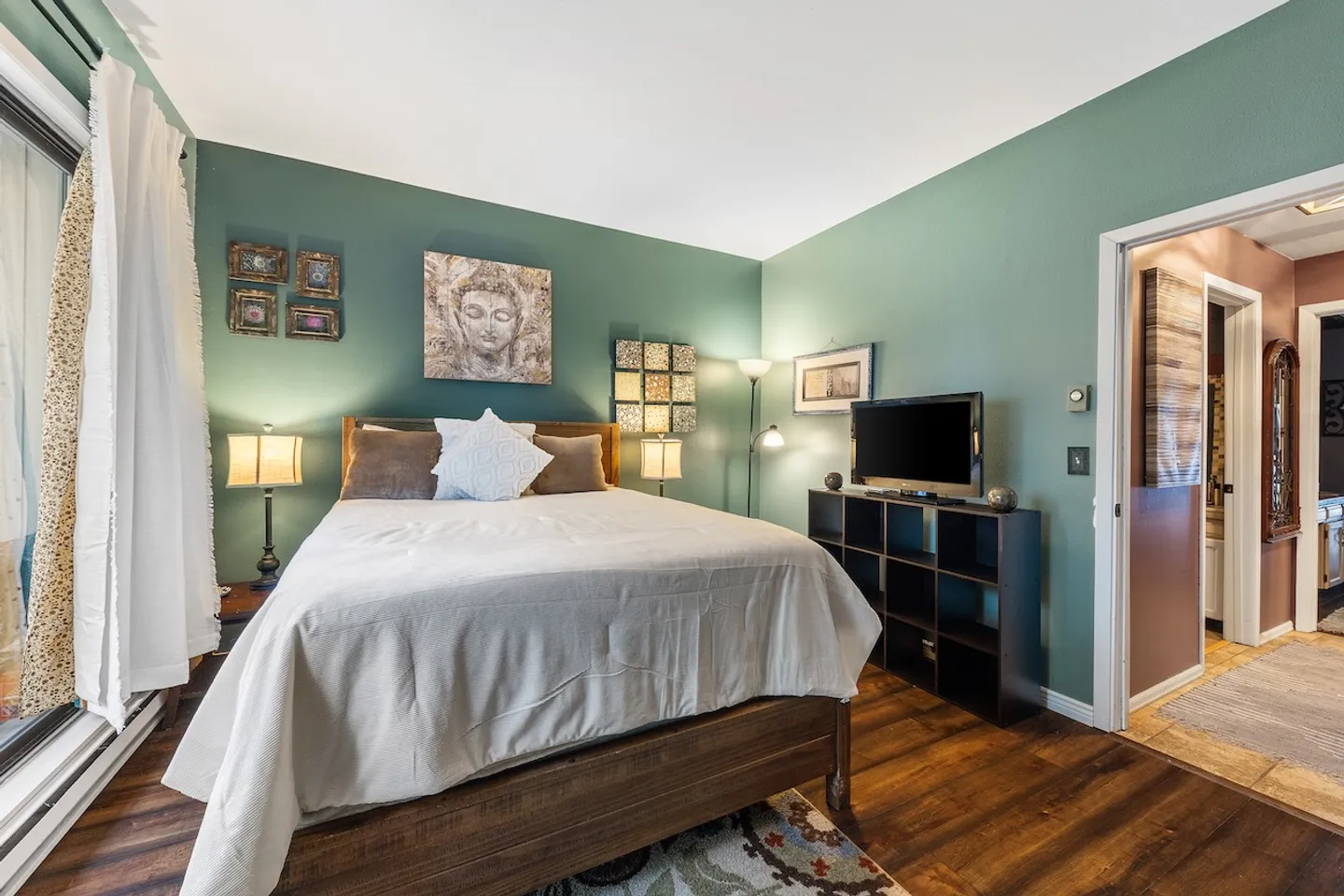 Bedroom with sage green walls, a bed made with light bedding, nightstands with lamps, a television on a shelving unit, and a doorway opening to an adjacent room.