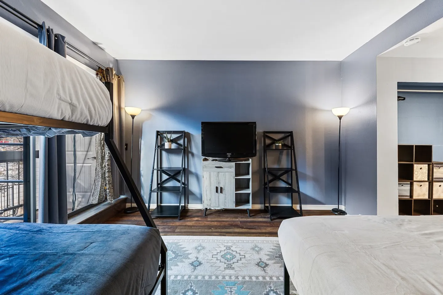 Bedroom featuring a blue accent wall, a television atop a small cabinet, open shelving units, floor lamps, and beds.