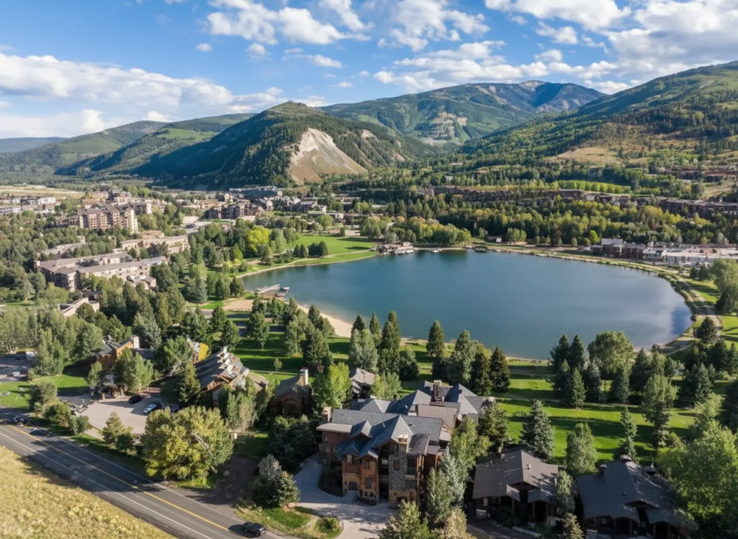Aerial view of a mountain resort town featuring large homes, a lake with a sandy beach area, and mountains under a blue sky.
