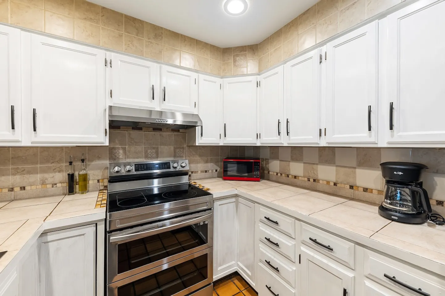 Kitchen corner featuring white cabinetry, tiled countertops, a stainless steel electric range, and small countertop appliances.