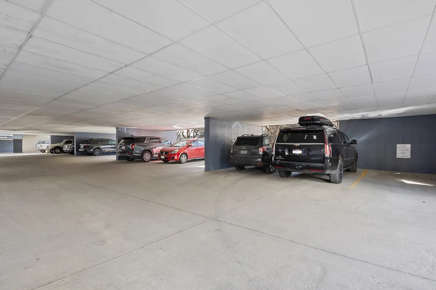 Covered parking garage with concrete flooring, low ceiling tiles, and parked cars.