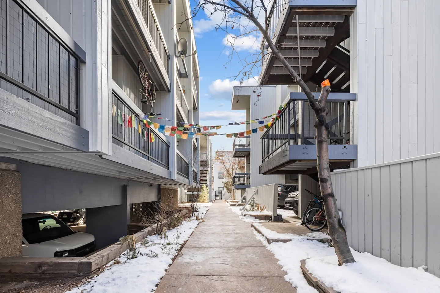 Exterior walkway between multi-story apartment buildings with snow patches and prayer flags overhead.