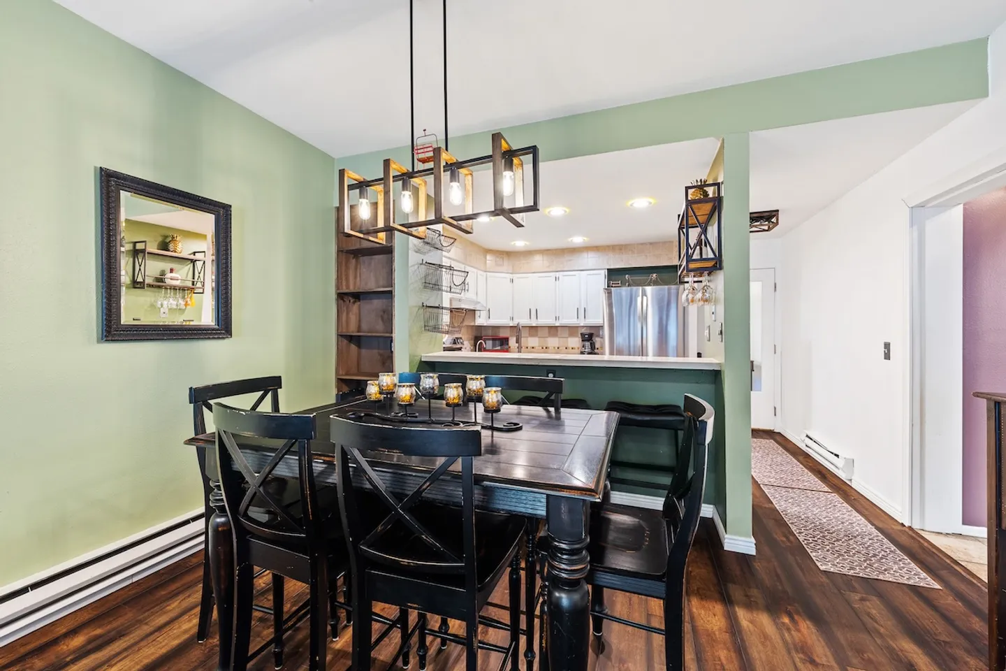 Dining area with a black wooden table and chairs under a modern chandelier, adjacent to a kitchen counter and hallway.