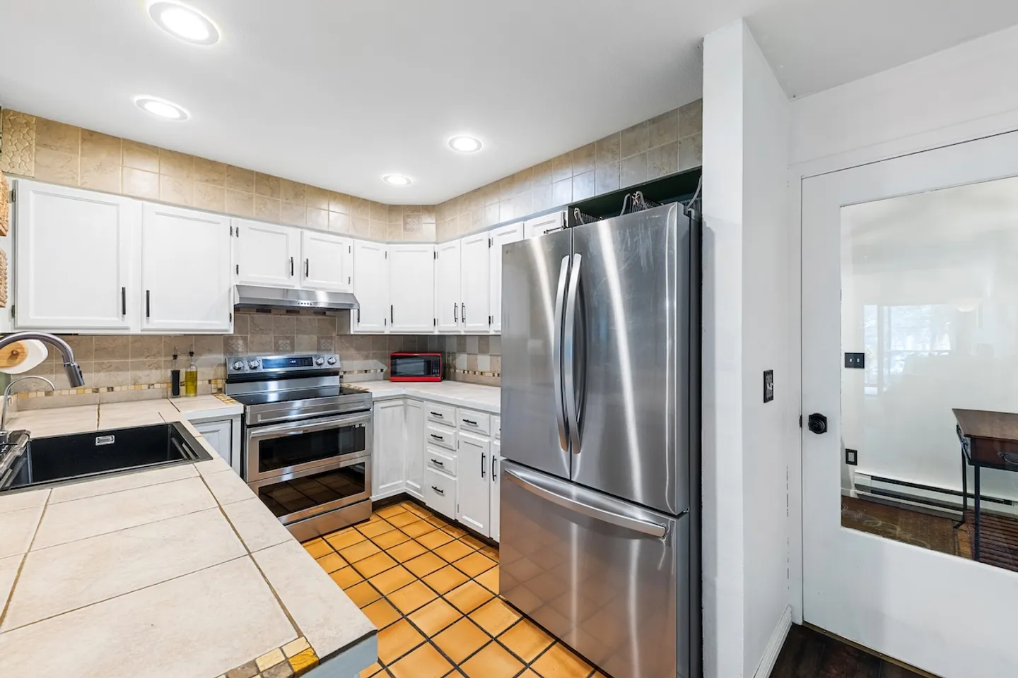Kitchen with white cabinetry, tile countertops, stainless steel appliances, and terracotta tile flooring.