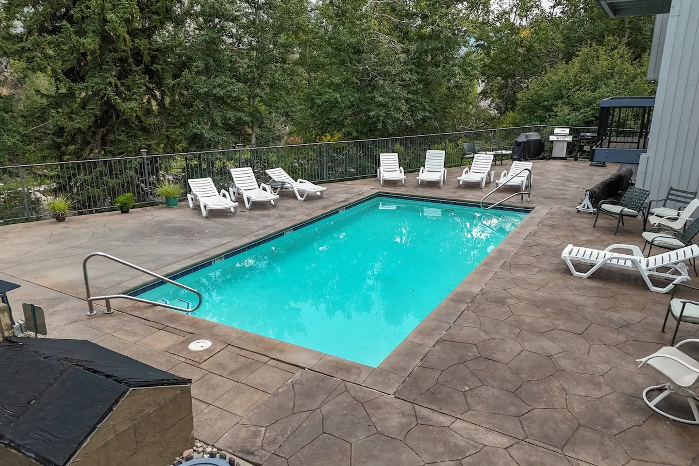 Outdoor swimming pool surrounded by a stamped concrete deck with white lounge chairs, enclosed by black metal fencing with trees in the background.