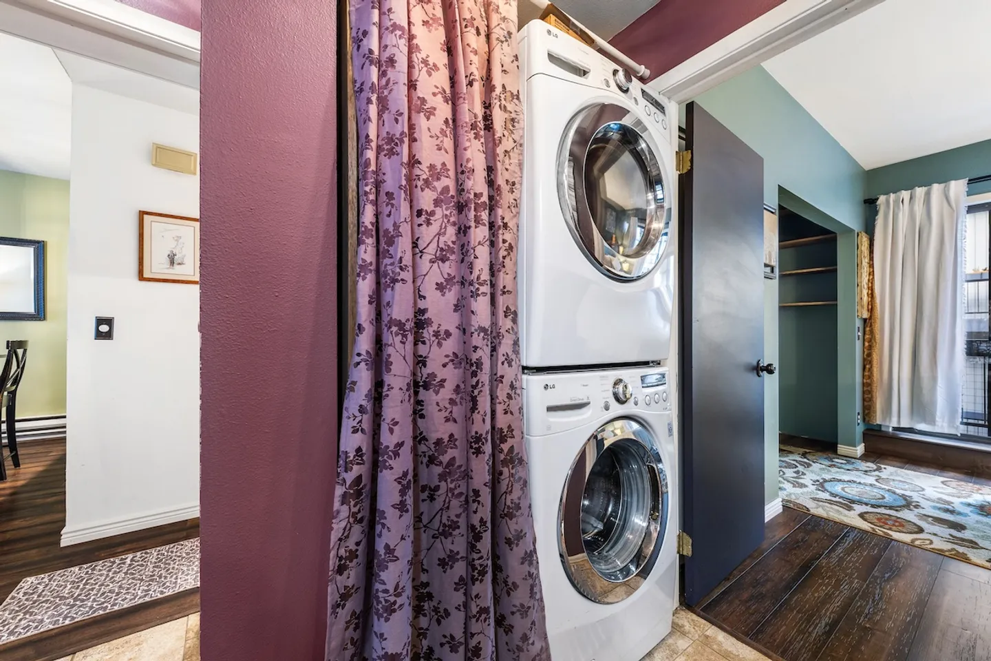 A stacked white front-load washer and dryer tucked in an alcove behind a patterned purple curtain between two rooms.