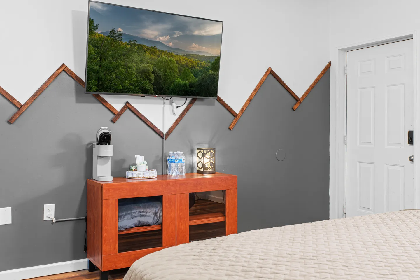 Bedroom corner featuring a wall-mounted TV over a small wooden cabinet, set against a two-tone painted wall with mountain-shaped wood trim.