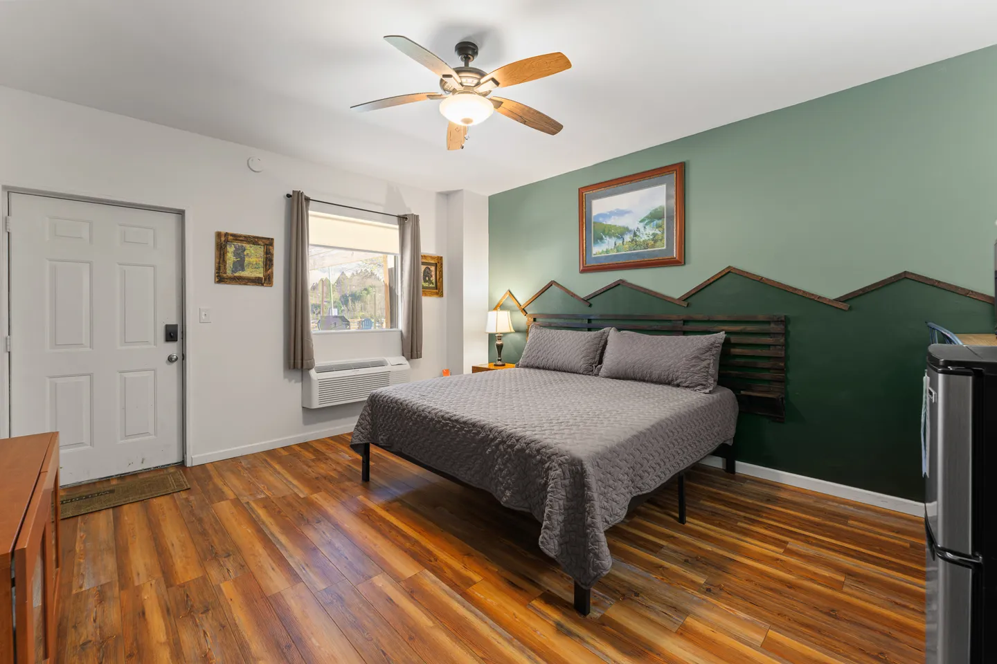 Bedroom featuring wood-style flooring, a bed with a dark gray quilt, an accent wall painted green with a mountain motif, and a ceiling fan.