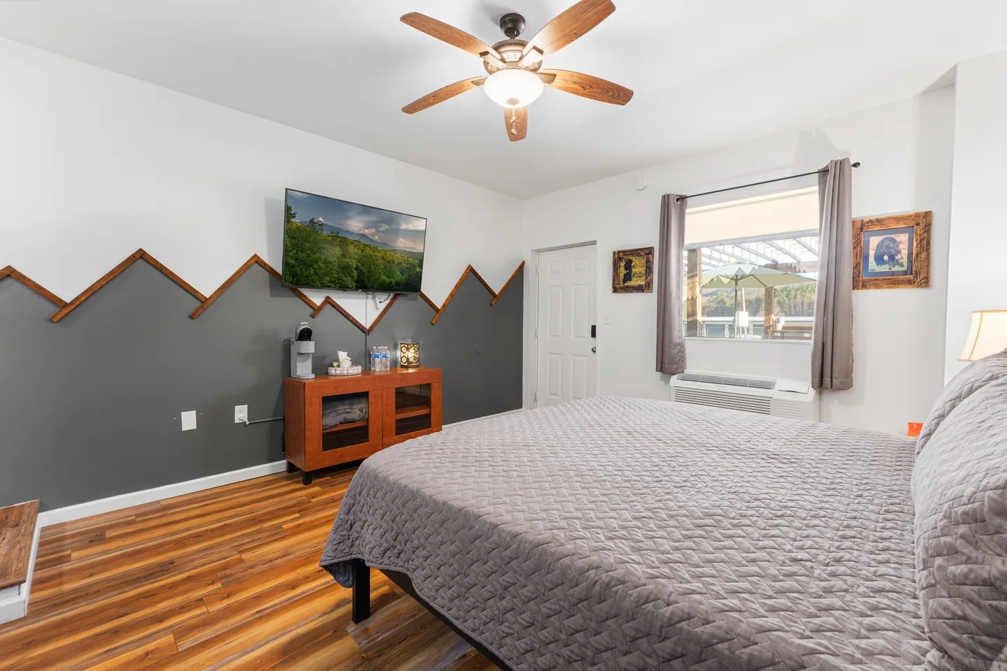 Bedroom with a bed made up with a gray quilt, wood-look flooring, ceiling fan, wall-mounted television, and a window.